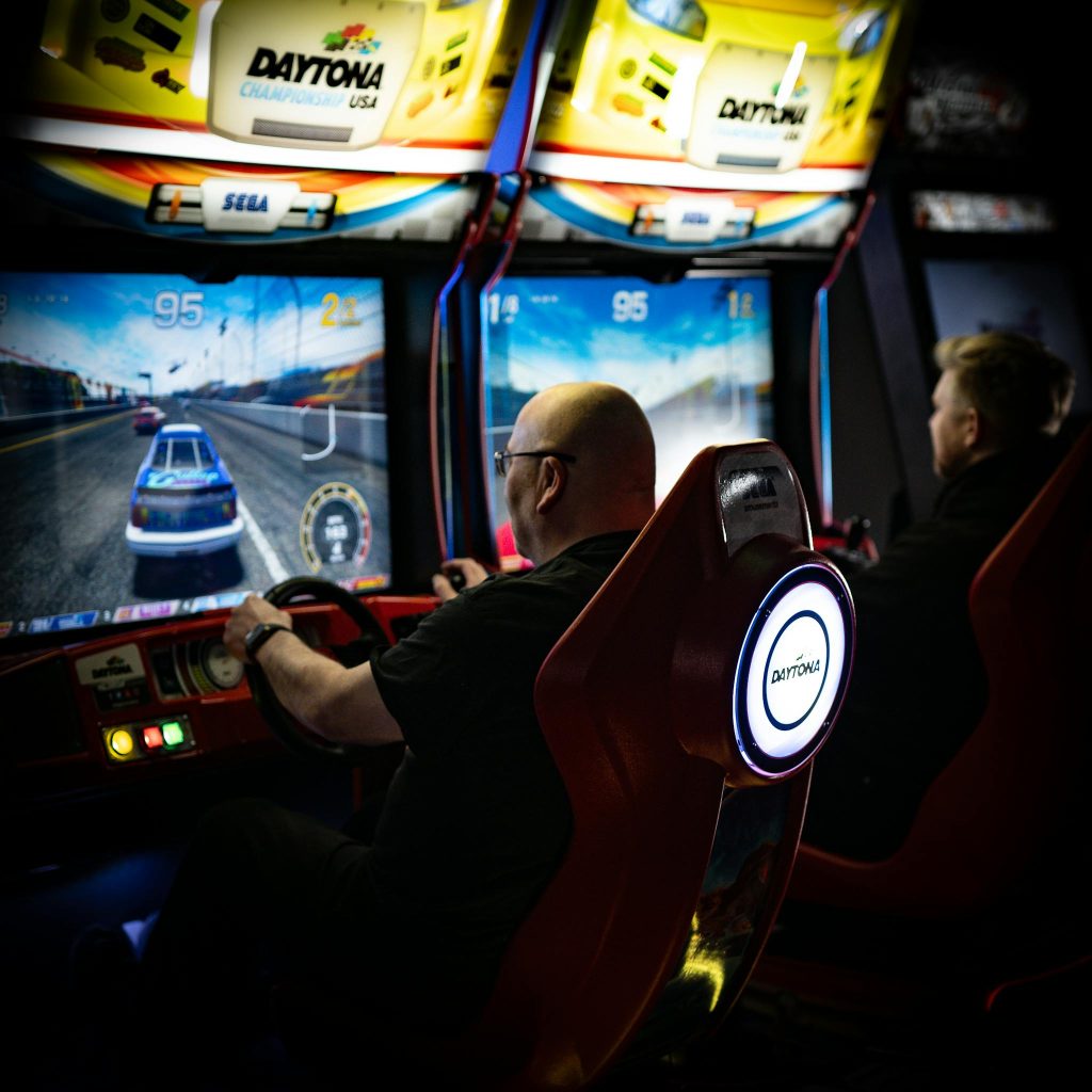 Two men immersed in Daytona racing arcade games at an indoor arcade.
